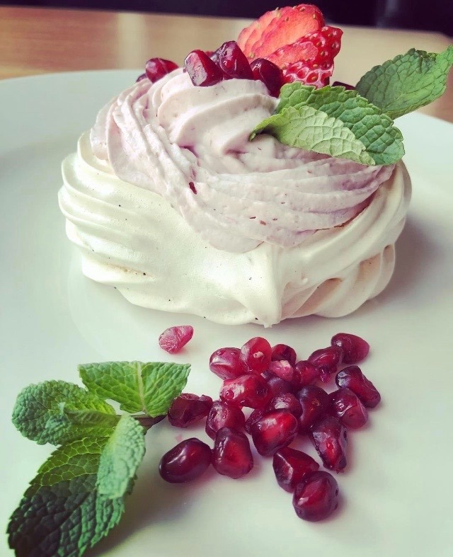 Delikatna beza Pavlova z kremem o różowym odcieniu, udekorowana świeżą truskawką, granatem i gałązkami mięty na białym talerzu.