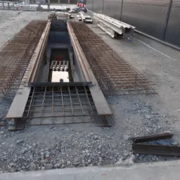 Przygotowanie zbrojenia do wylania fundamentów pod budynek przemysłowy, widoczne stalowe profile i siatka zbrojeniowa nad wykopem, w tle elementy konstrukcyjne i budynek z blachy trapezowej.