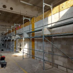 Wnętrze w trakcie budowy z widocznymi ścianami z betonu komórkowego, metalowymi rusztowaniami, drewnianymi elementami szalunkowymi i narzędziami budowlanymi na podłodze.