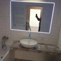 Biała łazienka z umywalką nablatową na blacie z patchworkowych płytek, lustro z podświetleniem LED, widoczna drabina w odbiciu lustra.