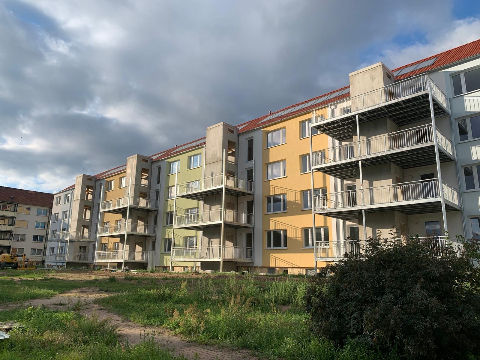 Nowoczesny budynek wielorodzinny z balkonami o stalowej konstrukcji, widoczne elementy ocynkowane, gotowe do montażu, fasada w odcieniach żółci i zieleni, na pierwszym planie zieleń.
