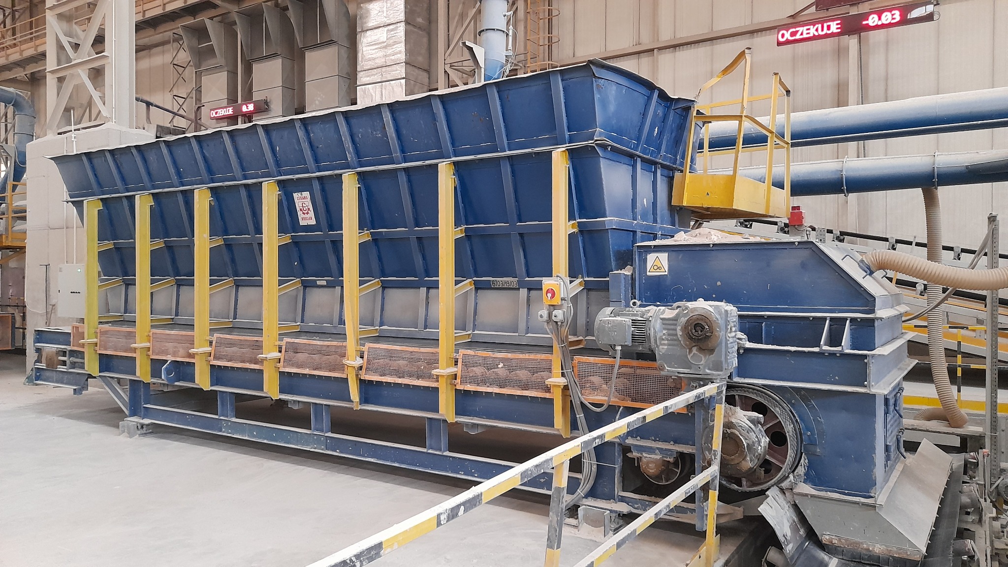 Duży, niebieski podajnik przemysłowy z żółtymi wzmocnieniami, znajdujący się w hali produkcyjnej, z widocznymi silnikami i elementami przenośnika.