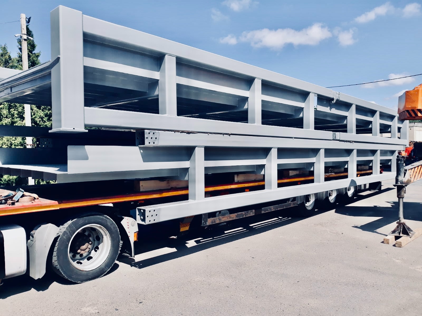 Szara, stalowa konstrukcja hali w trakcie transportu na naczepie ciężarówki, zabezpieczona drewnianymi belkami, widoczne elementy żurawia.