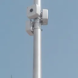 Srebrny słup z zamontowanymi trzema białymi kamerami monitoringu na zewnątrz, na tle błękitnego nieba i fragmentu zielonego drzewa.