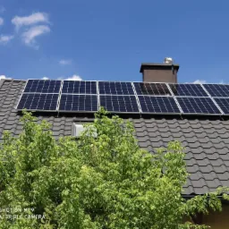 Panele fotowoltaiczne na dachu z szarej dachówki, częściowo zasłonięte przez zielone drzewo, widoczne kominy i błękitne niebo