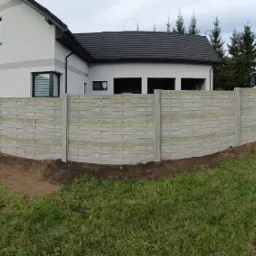 Długi, szary płot z prefabrykatów betonowych o fakturze imitującej plecionkę, oddzielający posesję z nowoczesnym domem od sąsiednich działek; widoczny wykop wzdłuż ogrodzenia i sprzęt budowlany w tle.