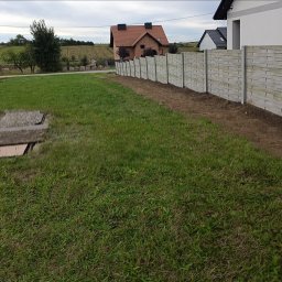 FHU Wool-Max - Długi, szary płot z prefabrykowanych elementów imitujących drewno oddziela trawnik od sąsiedniej działki. Świeżo wykopana ziemia wzdłuż ogrodzenia. W tle domy i pole.