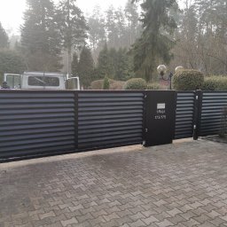 FHU Wool-Max - Nowoczesna, przesuwna brama w kolorze antracytu z poziomymi panelami, z adresem '1 Maja 173/175'. W tle zieleń drzew i krzewów, częściowo widoczny samochód dostawczy.