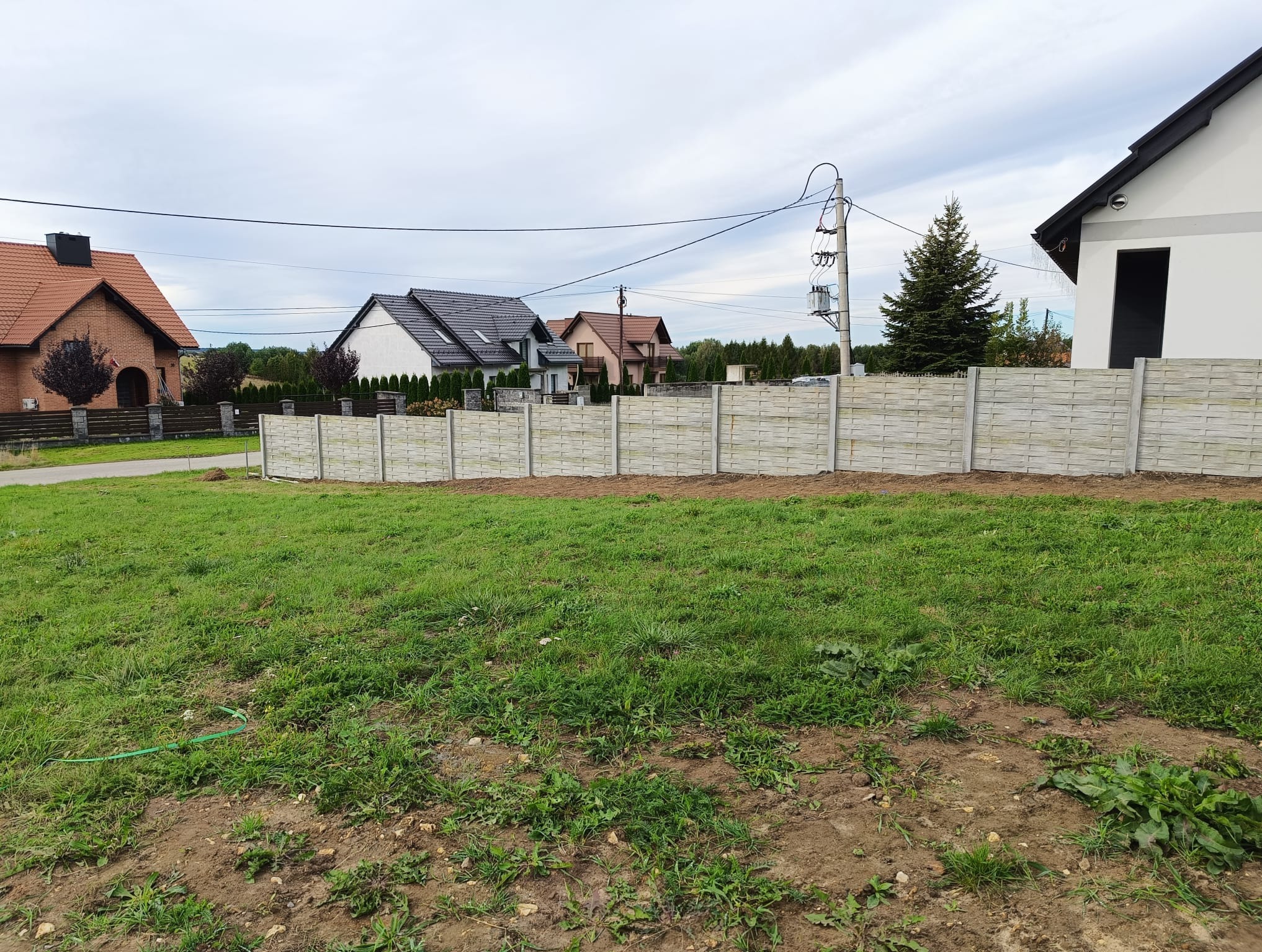 Nowo postawione, ażurowe ogrodzenie z prefabrykatów betonowych, oddzielające trawnik od zabudowy jednorodzinnej w tle, widok w pochmurny dzień.