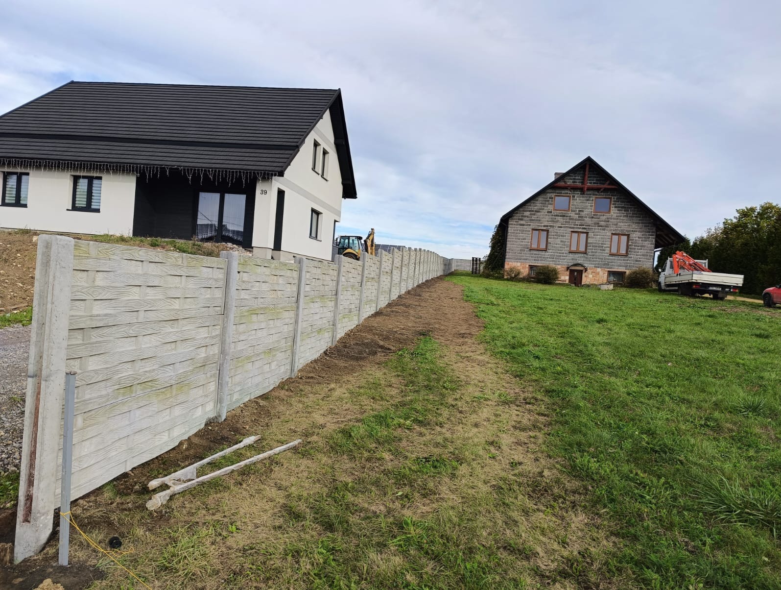 Długi, szary, betonowy płot panelowy oddzielający dwie posesje z domami w różnym stylu architektonicznym, z widocznym sprzętem budowlanym w tle.