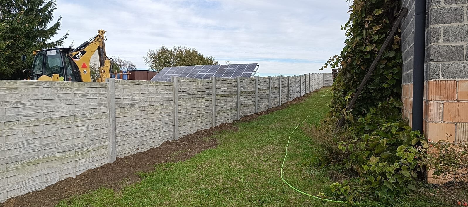 Długi, szary, betonowy płot o wzorze plecionki, ciągnący się wzdłuż trawnika. W tle widoczny żółty traktor, panele słoneczne i budynek z cegły.