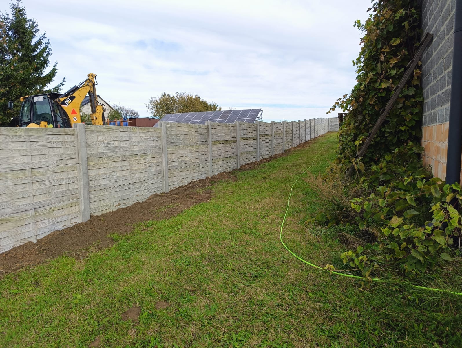 Długi, szary płot z betonowych paneli w trakcie montażu, widoczna świeżo przekopana ziemia i maszyna budowlana CAT w tle, panele słabo widoczne z fakturą imitującą drewno.