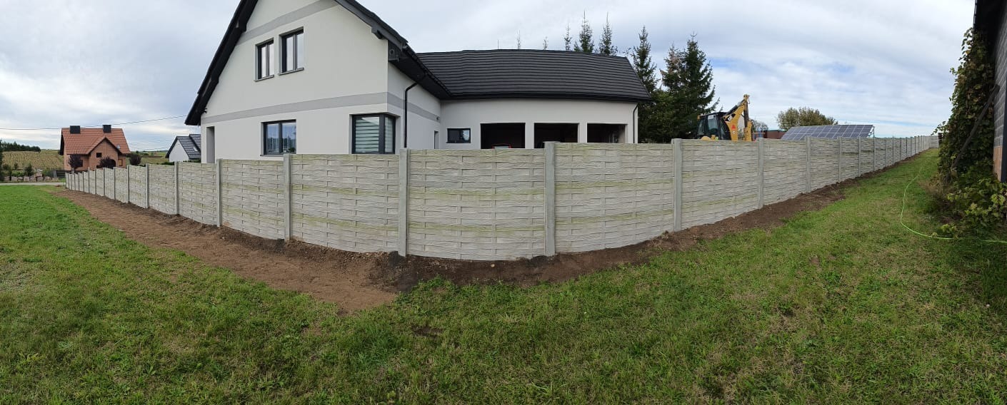 Długi, szary płot z prefabrykatów betonowych o fakturze imitującej plecionkę, oddzielający posesję z nowoczesnym domem od sąsiednich działek; widoczny wykop wzdłuż ogrodzenia i sprzęt budowlany w tle.