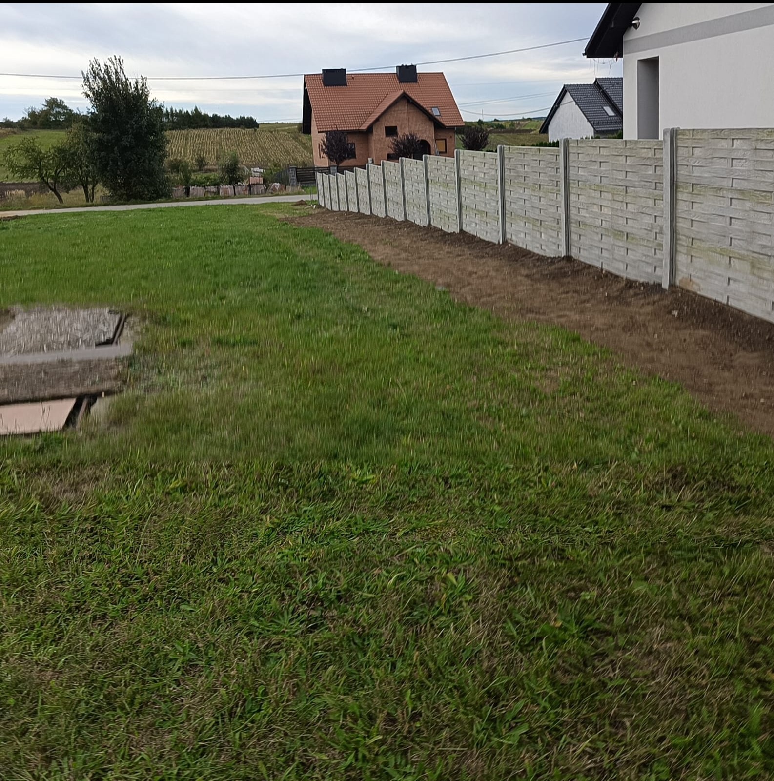 Długi, szary płot z prefabrykowanych elementów imitujących drewno oddziela trawnik od sąsiedniej działki. Świeżo wykopana ziemia wzdłuż ogrodzenia. W tle domy i pole.
