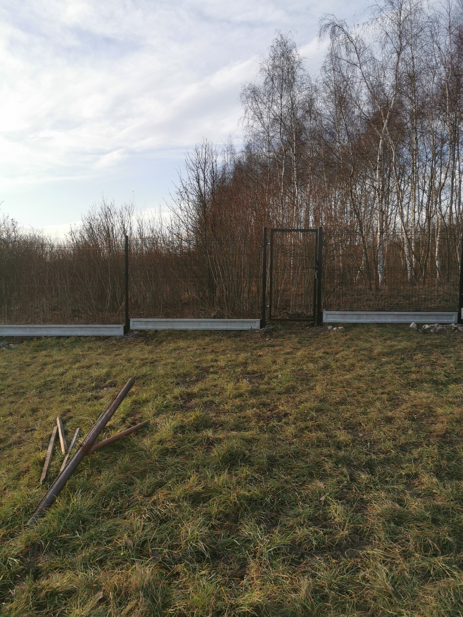 Fragment montowanego ogrodzenia panelowego z bramką na nierównym terenie. Widoczne betonowe podmurówki, słupki i elementy siatki. W tle drzewa i krzewy.