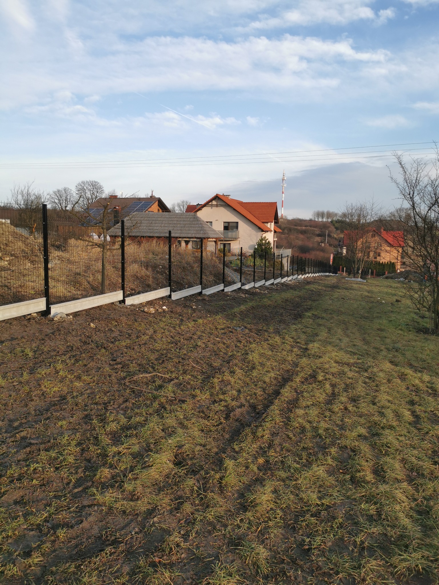 Długi, panelowy płot z podmurówką, ciągnący się wzdłuż nierównego terenu. W tle domy z czerwonymi dachami i błękitne niebo z chmurami.