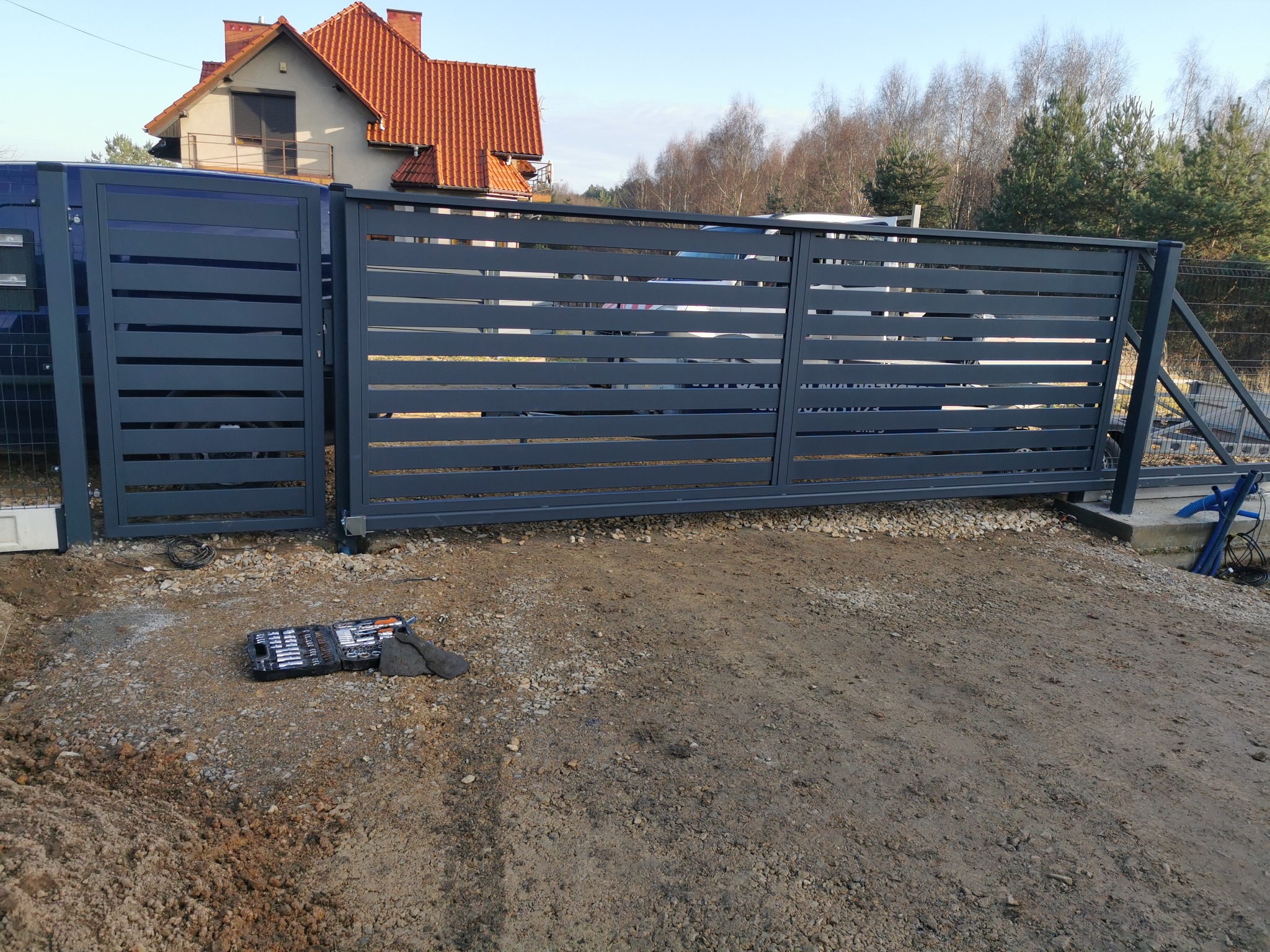 Nowoczesna, przesuwna brama w kolorze grafitowym z poziomymi panelami, zamontowana na posesji. Na ziemi widoczny zestaw narzędzi.
