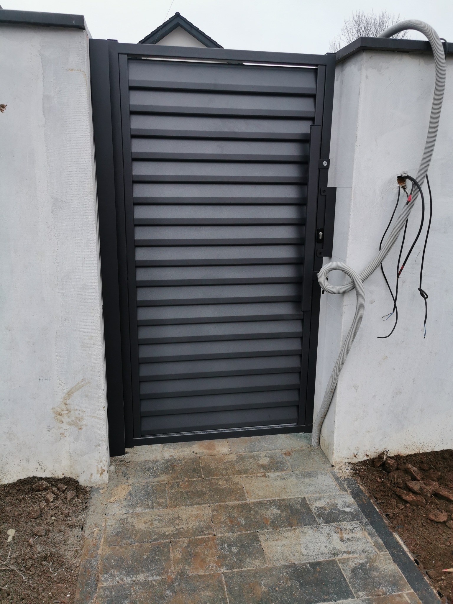 Nowoczesna, grafitowa brama wjazdowa z poziomych paneli, zamontowana w surowym, białym murze. Widoczne przewody elektryczne i kostka brukowa.