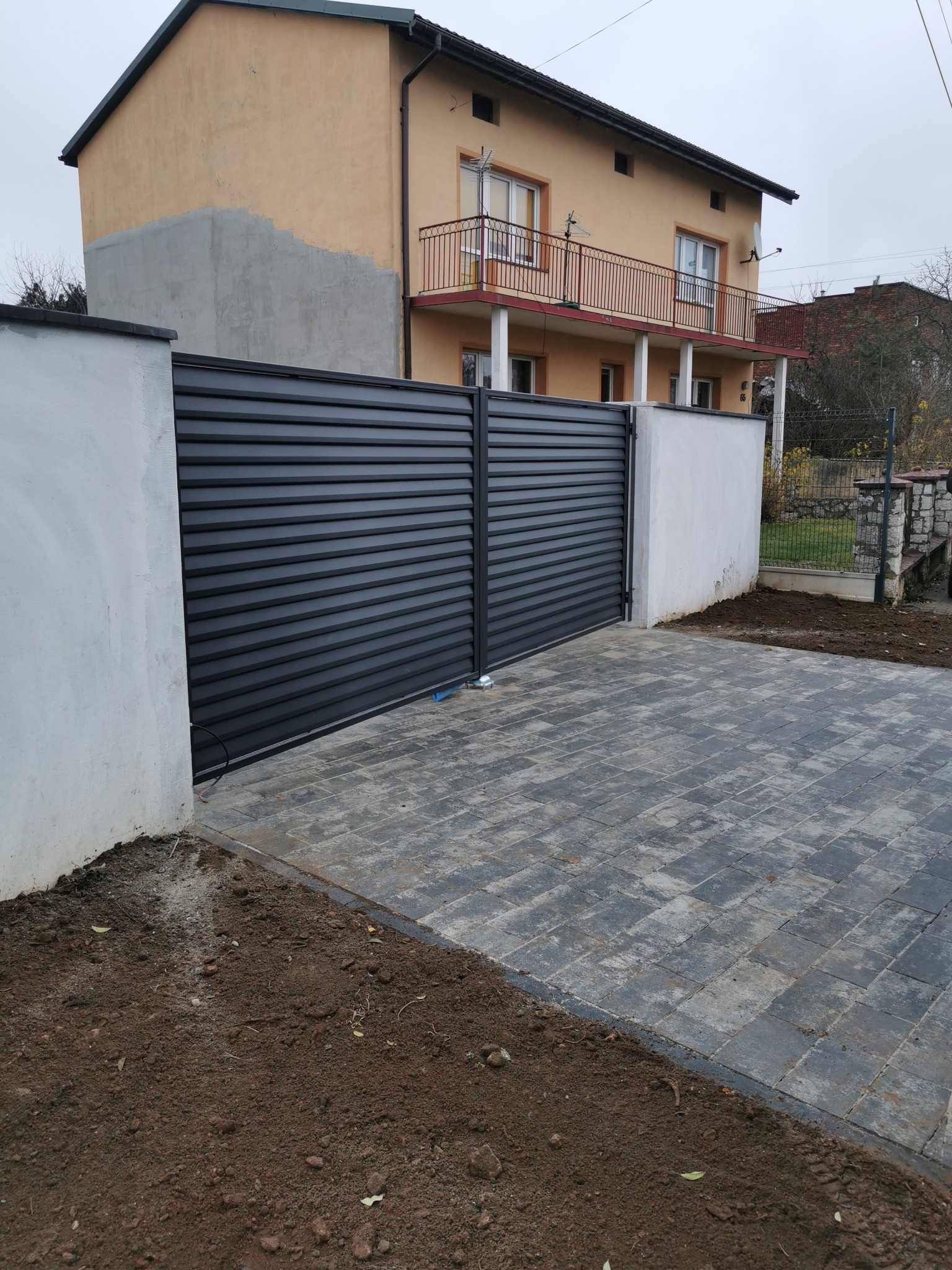 Nowoczesna, pozioma brama wjazdowa w kolorze antracytu, osadzona między białymi słupkami. Kostka brukowa prowadzi do bramy. W tle dom z balkonem.