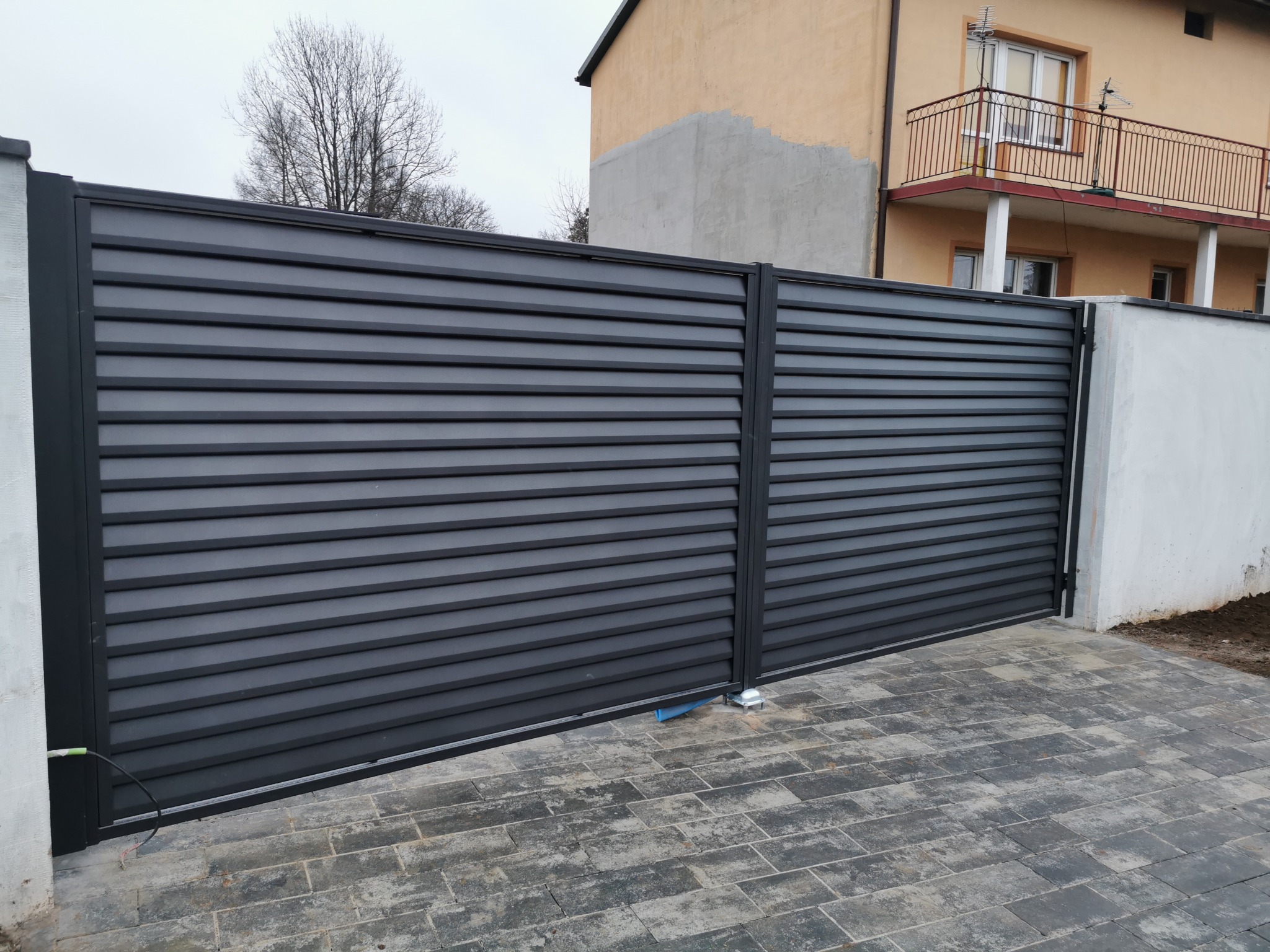 Nowoczesna, pozioma brama przesuwna z metalowymi lamelami w kolorze grafitowym, zamontowana w Kluczach. Widoczna kostka brukowa i fragment budynku w tle.