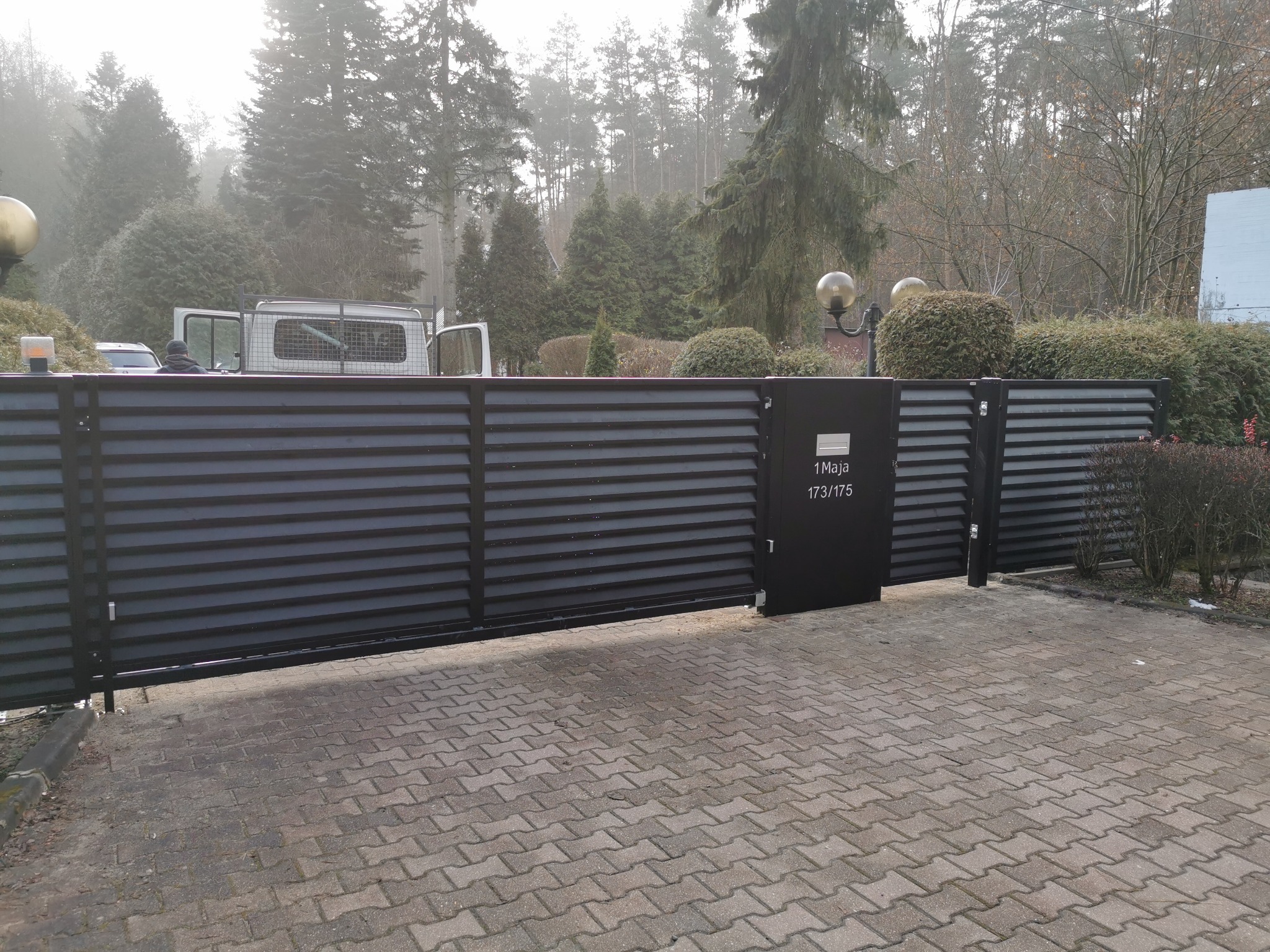Nowoczesna, przesuwna brama w kolorze antracytu z poziomymi panelami, z adresem '1 Maja 173/175'. W tle zieleń drzew i krzewów, częściowo widoczny samochód dostawczy.