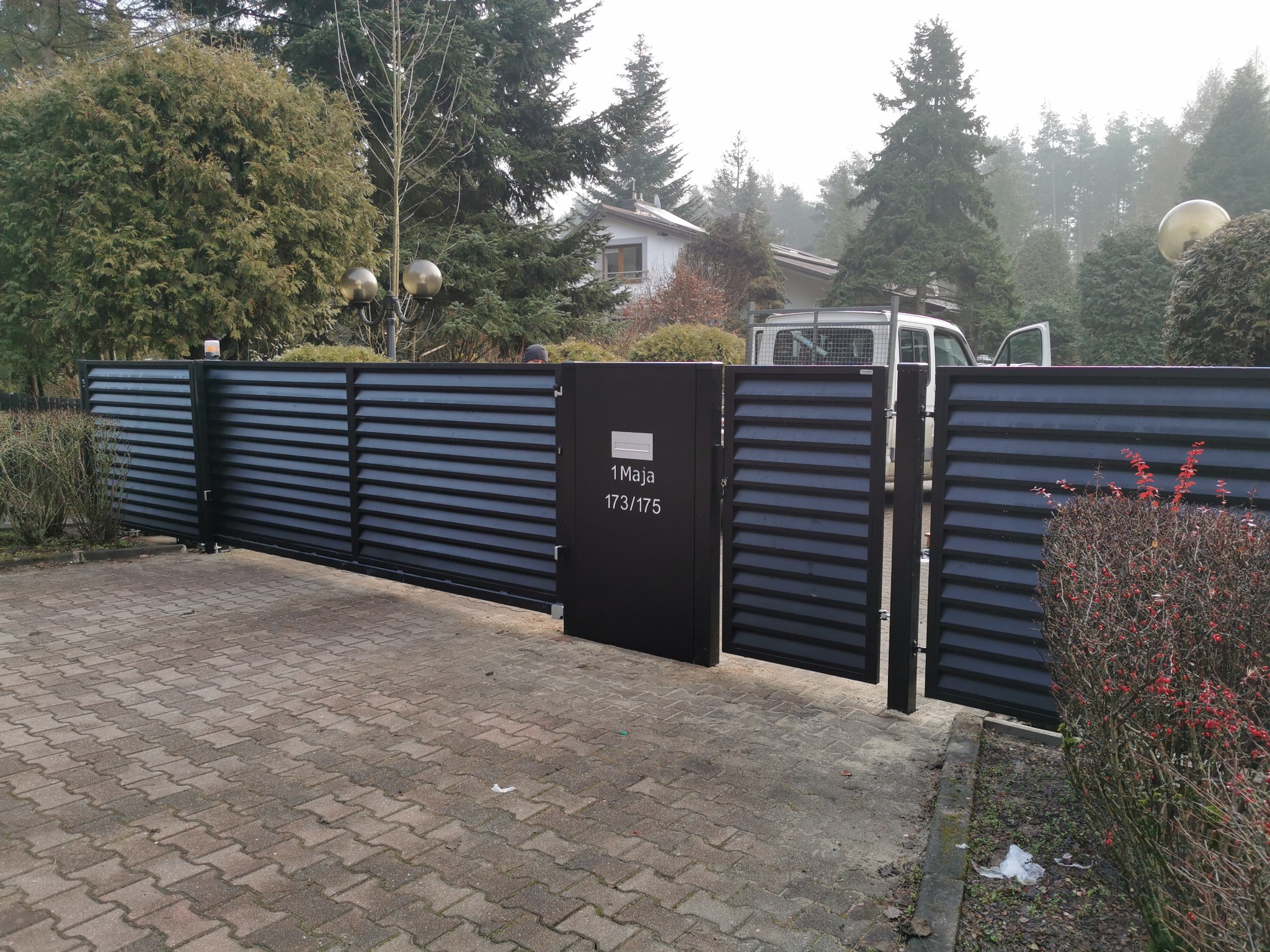 Nowoczesna, pozioma brama przesuwna w kolorze antracytowym z furtką, z numerem domu '1 Maja 173/175', na tle zieleni i domu. Widok z zewnątrz posesji.