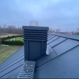 Zakład Dekarski Piotr Bystranowski - Szary dach z blachy na rąbek stojący z zamontowanym stopniem kominiarskim i kominem, widok na posesję z trawnikiem i żywopłotem w pochmurny dzień.