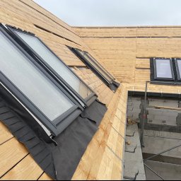 Zakład Dekarski Piotr Bystranowski - Montaż okien dachowych w drewnianej konstrukcji dachu, widoczne elementy izolacji i rusztowanie przy ścianie budynku.