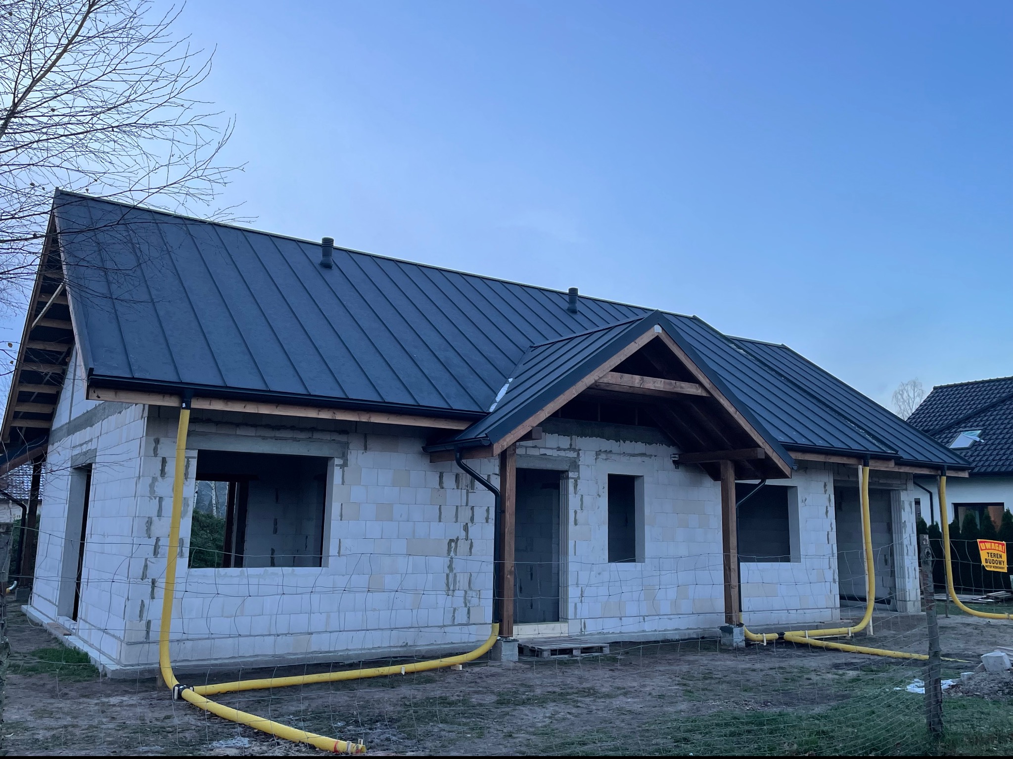 Dom w trakcie budowy z ciemnym, nowym dachem z blachy na rąbek stojący, widoczne żółte węże wentylacyjne doprowadzone do budynku, ściany z białych bloczków, brak okien i drzwi, teren budowy...
