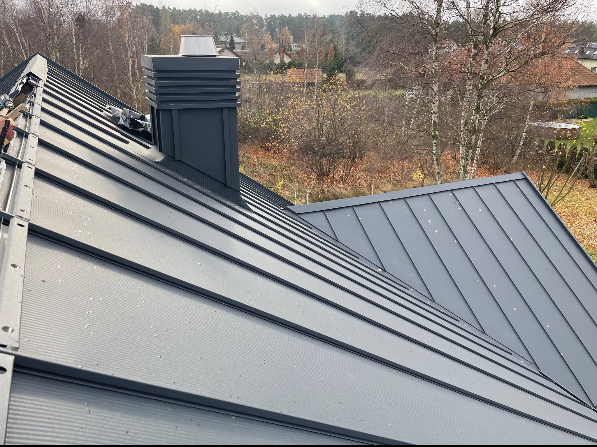 Nowy, grafitowy dach z blachy na rąbek stojący, z widocznym kominem i elementami montażowymi, w tle jesienny krajobraz z drzewami.