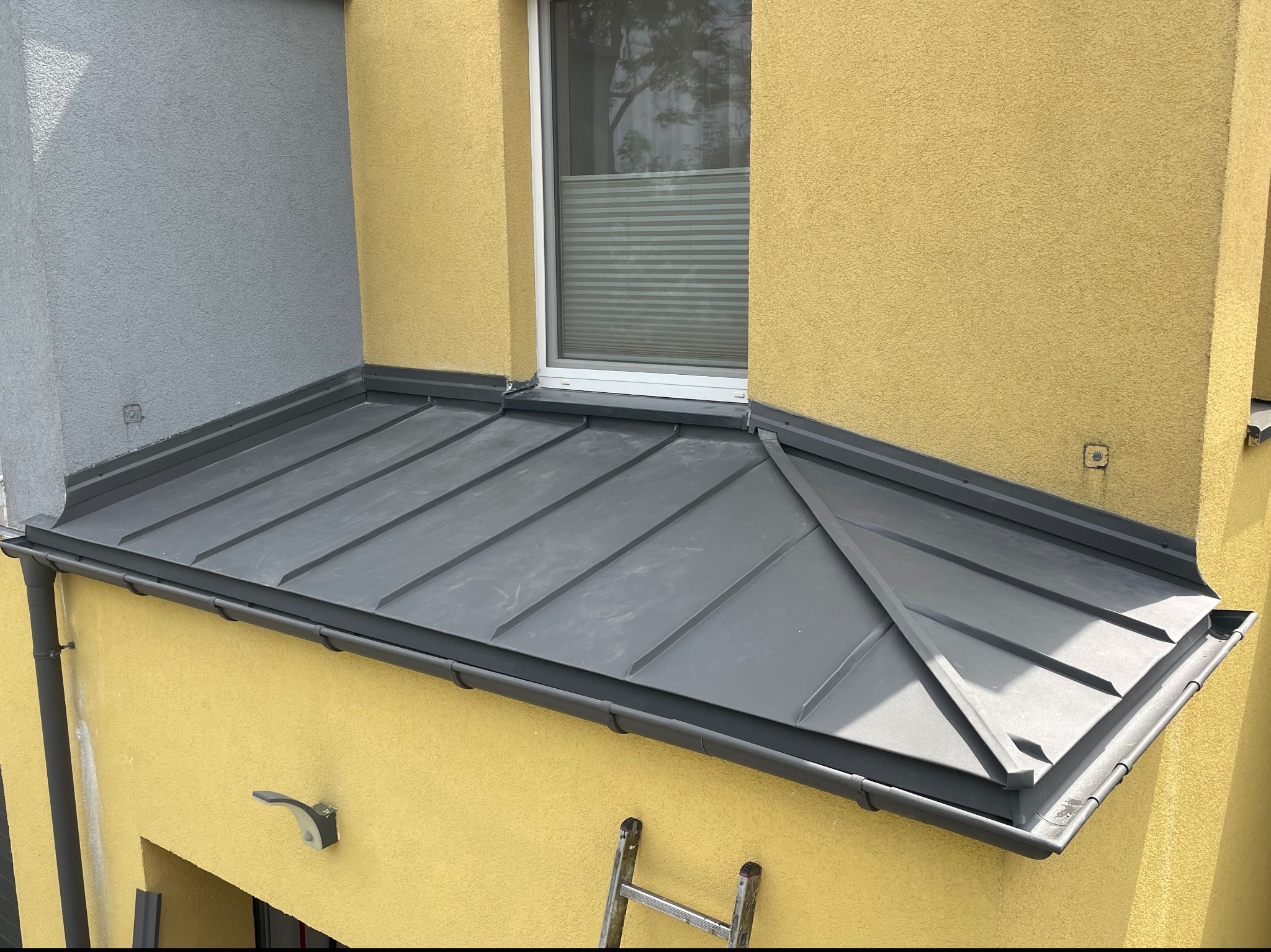 Nowoczesny dach z blachy na budynku o żółtej i szarej elewacji, widoczne okno z żaluzjami plisowanymi oraz rynna, fragment drabiny oparty o ścianę.