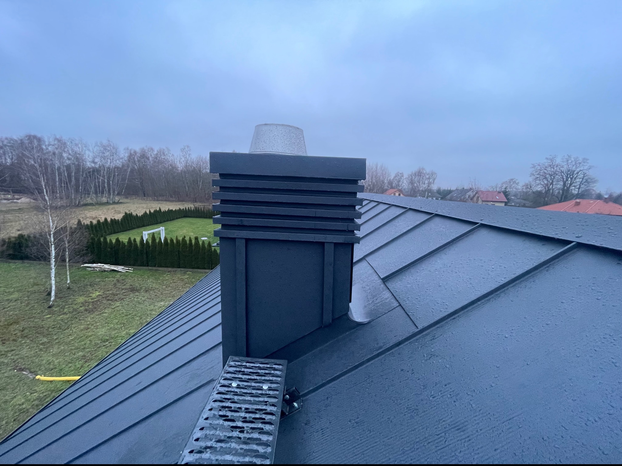 Szary dach z blachy na rąbek stojący z zamontowanym stopniem kominiarskim i kominem, widok na posesję z trawnikiem i żywopłotem w pochmurny dzień.