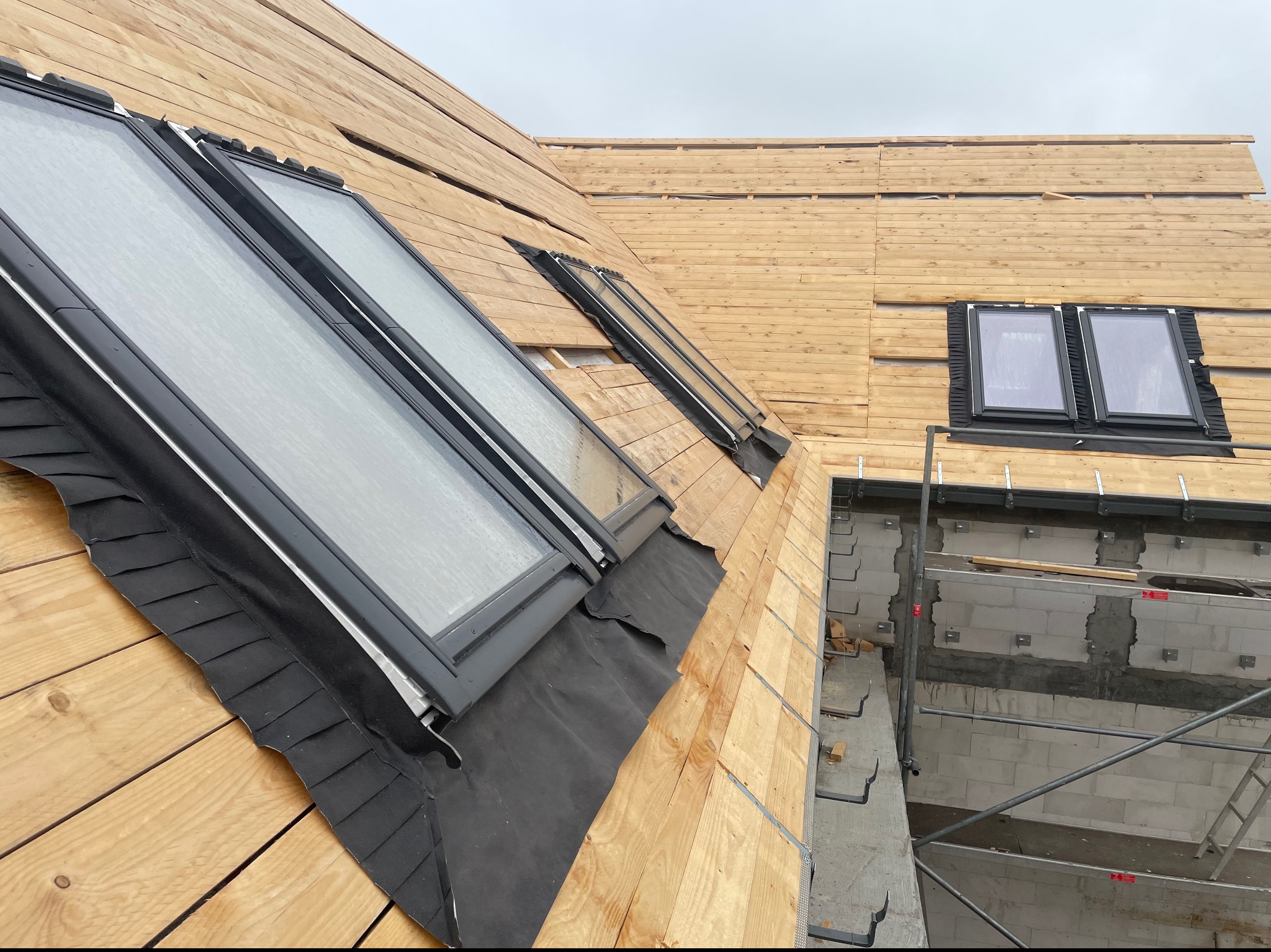 Montaż okien dachowych w drewnianej konstrukcji dachu, widoczne elementy izolacji i rusztowanie przy ścianie budynku.