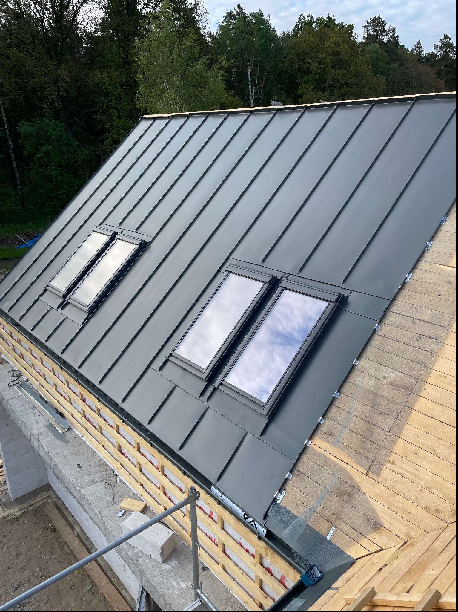 Nowy, grafitowy dach z blachy na rąbek stojący z zamontowanymi oknami dachowymi, widoczne elementy konstrukcji więźby dachowej i fragment murowanej ściany.