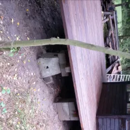 Konstrukcja tarasu z desek drewnianych, podparta na betonowych bloczkach, widoczny fragment drzewa i budynku w tle.