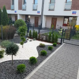 Nowoczesny ogr&oacute;d przed szeregowcem z białym i czarnym grysem, ozdobnymi drzewkami i krzewami, oraz szarą kostką brukową.