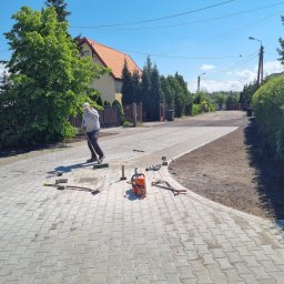 FIRMA REM-BUD2 Krzysztof Obrębski - Układanie kostki brukowej na posesji. Brukarz zamiata kostkę, obok leżą narzędzia: przecinarka, młotek. Widok na fragment ułożonej kostki i przygotowany teren.