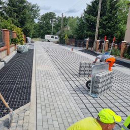 FIRMA REM-BUD2 Krzysztof Obrębski - Układanie jasnoszarej kostki brukowej na posesji. Widoczny fragment ułożonej nawierzchni, pracownicy z wózkiem pełnym kostki i narzędzia. W tle zabudowa jednorodzinna.