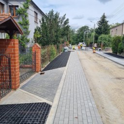 FIRMA REM-BUD2 Krzysztof Obrębski - Ułożona szara kostka brukowa na ulicy z kratką drenażową przy posesji. W tle pracownicy w żółtych kamizelkach i biały samochód dostawczy.