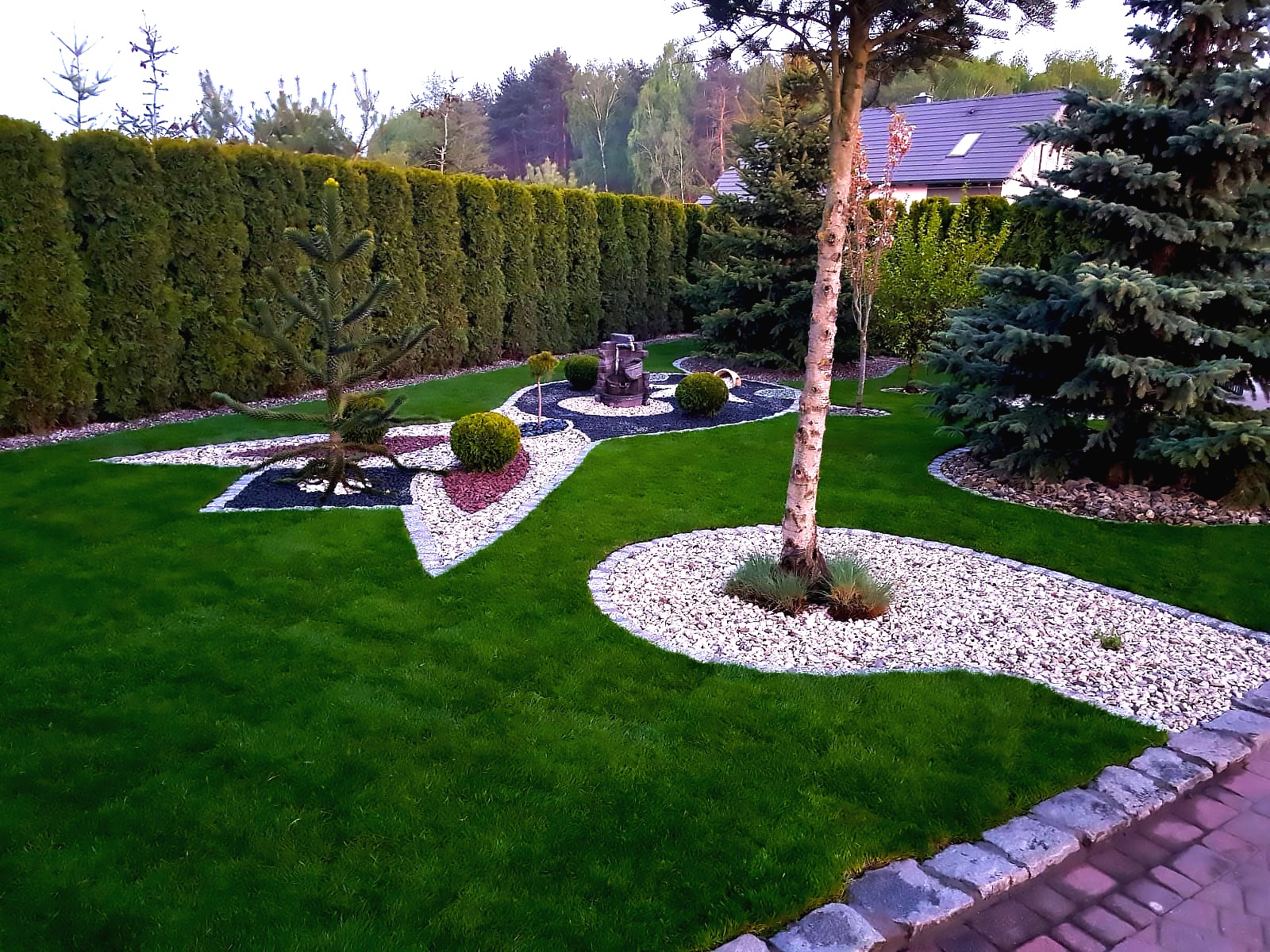 Estetyczny ogród z zielonym trawnikiem, ozdobnymi rabatami z kamieni i krzewów, oraz fontanną. Tuje w tle tworzą naturalny żywopłot.