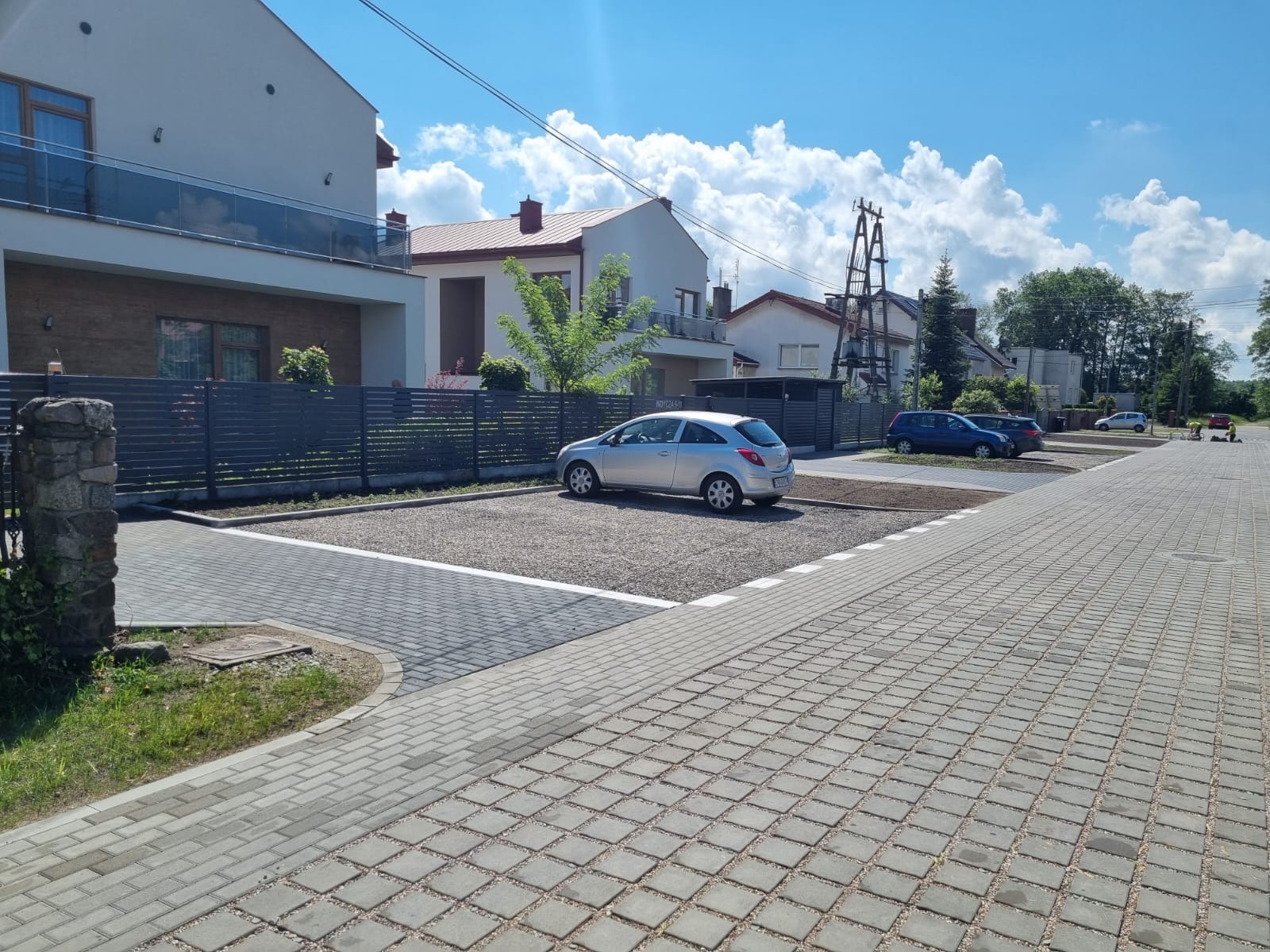 Wykończony parking z kostki brukowej i żwiru, z zaparkowanym srebrnym autem. Widok na domy w tle, słoneczny dzień. Estetyczne wykończenie.