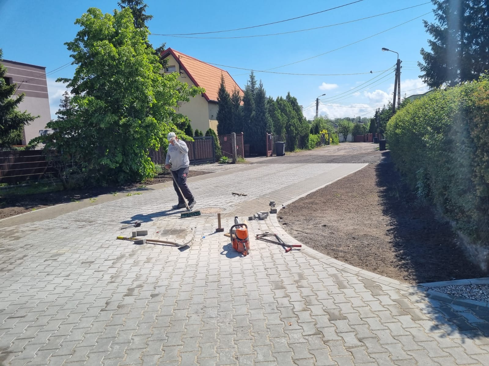 Układanie kostki brukowej na posesji. Brukarz zamiata kostkę, obok leżą narzędzia: przecinarka, młotek. Widok na fragment ułożonej kostki i przygotowany teren.