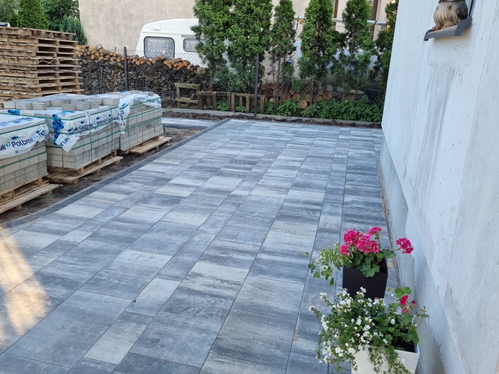 Ułożona szara kostka brukowa z paletami kostki w tle, obok ściany budynku z kwiatami w donicach. Widoczny fragment posesji z przyczepą kempingową.