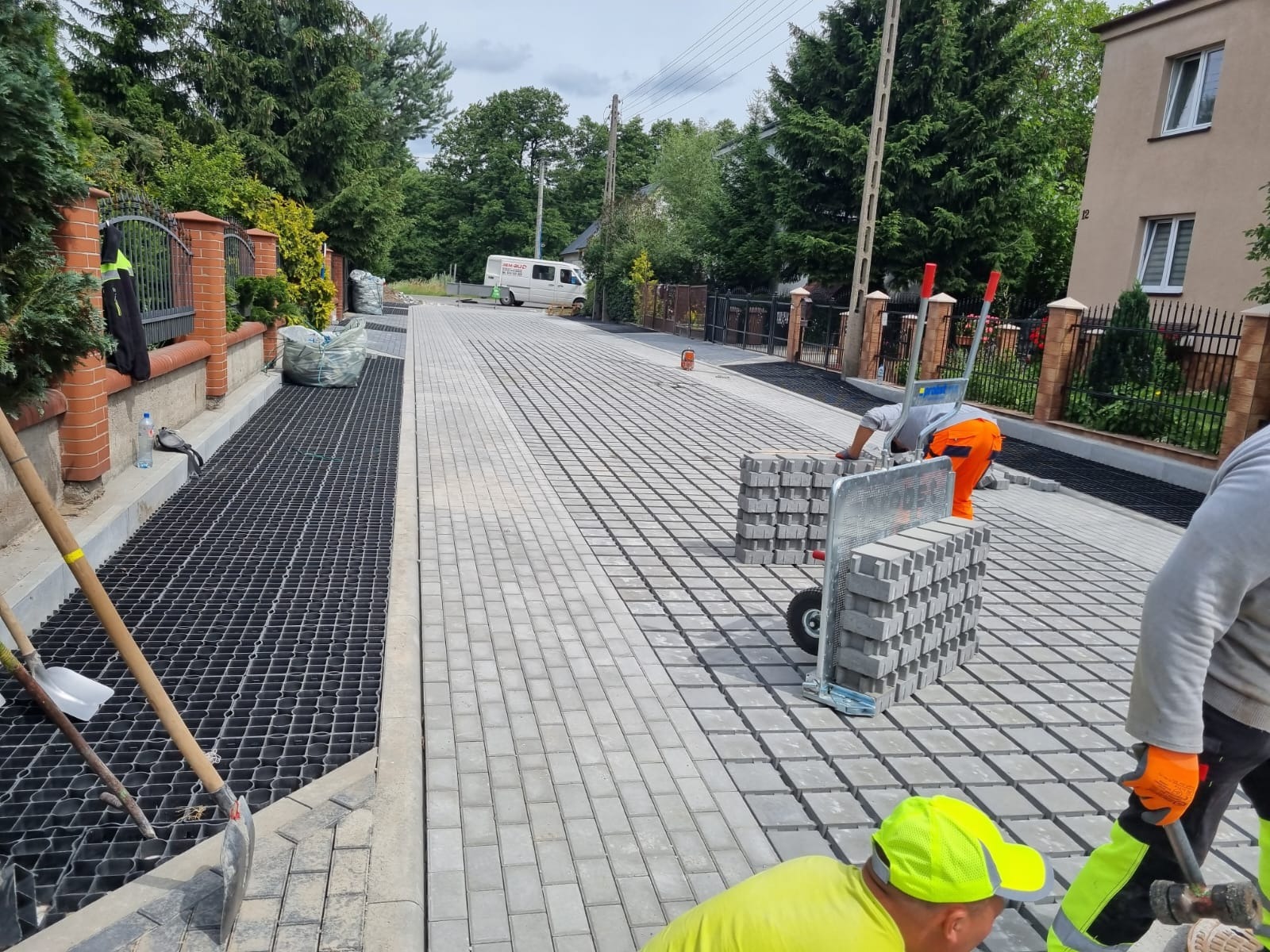 Układanie jasnoszarej kostki brukowej na posesji. Widoczny fragment ułożonej nawierzchni, pracownicy z wózkiem pełnym kostki i narzędzia. W tle zabudowa jednorodzinna.