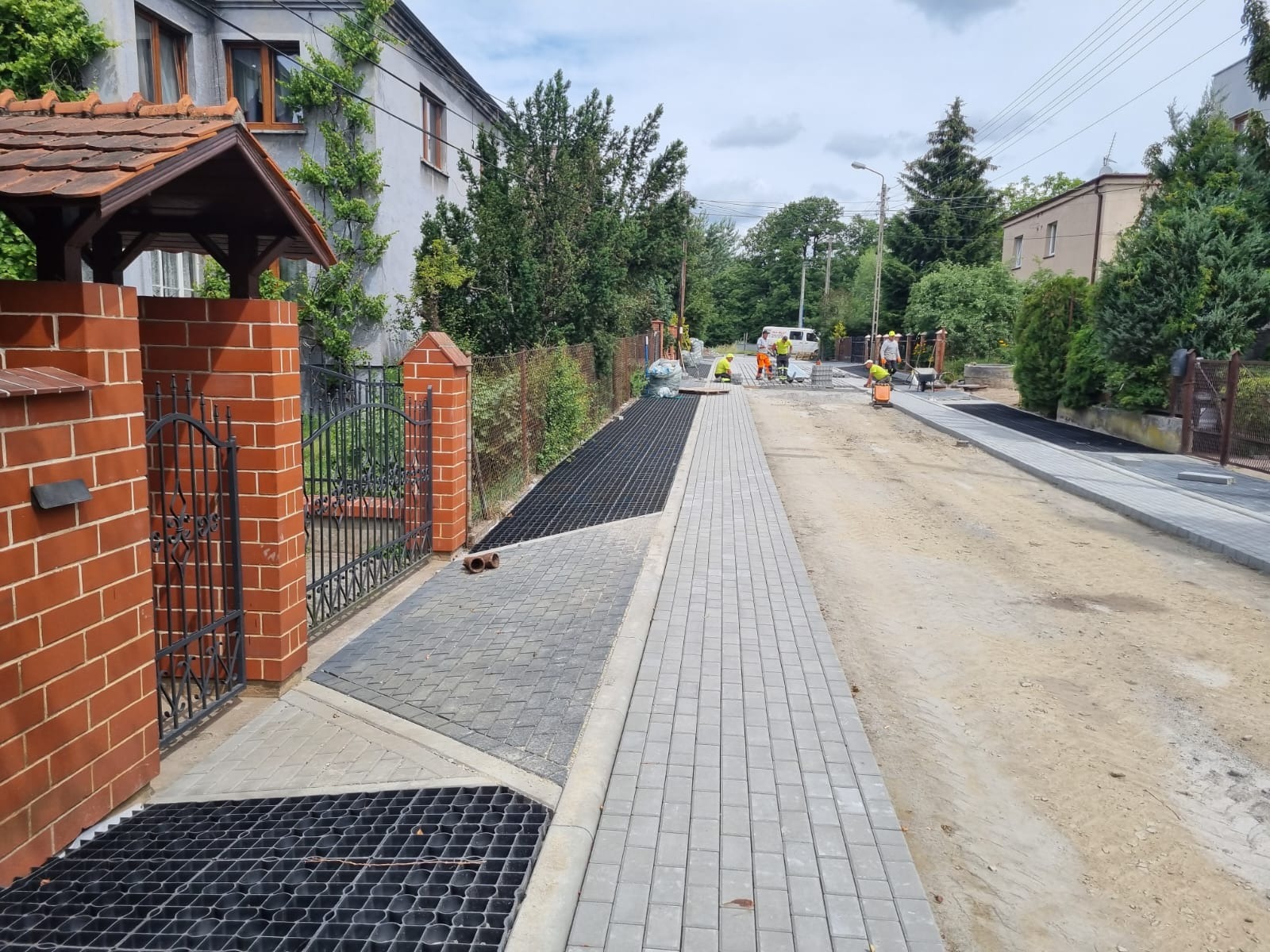 Ułożona szara kostka brukowa na ulicy z kratką drenażową przy posesji. W tle pracownicy w żółtych kamizelkach i biały samochód dostawczy.