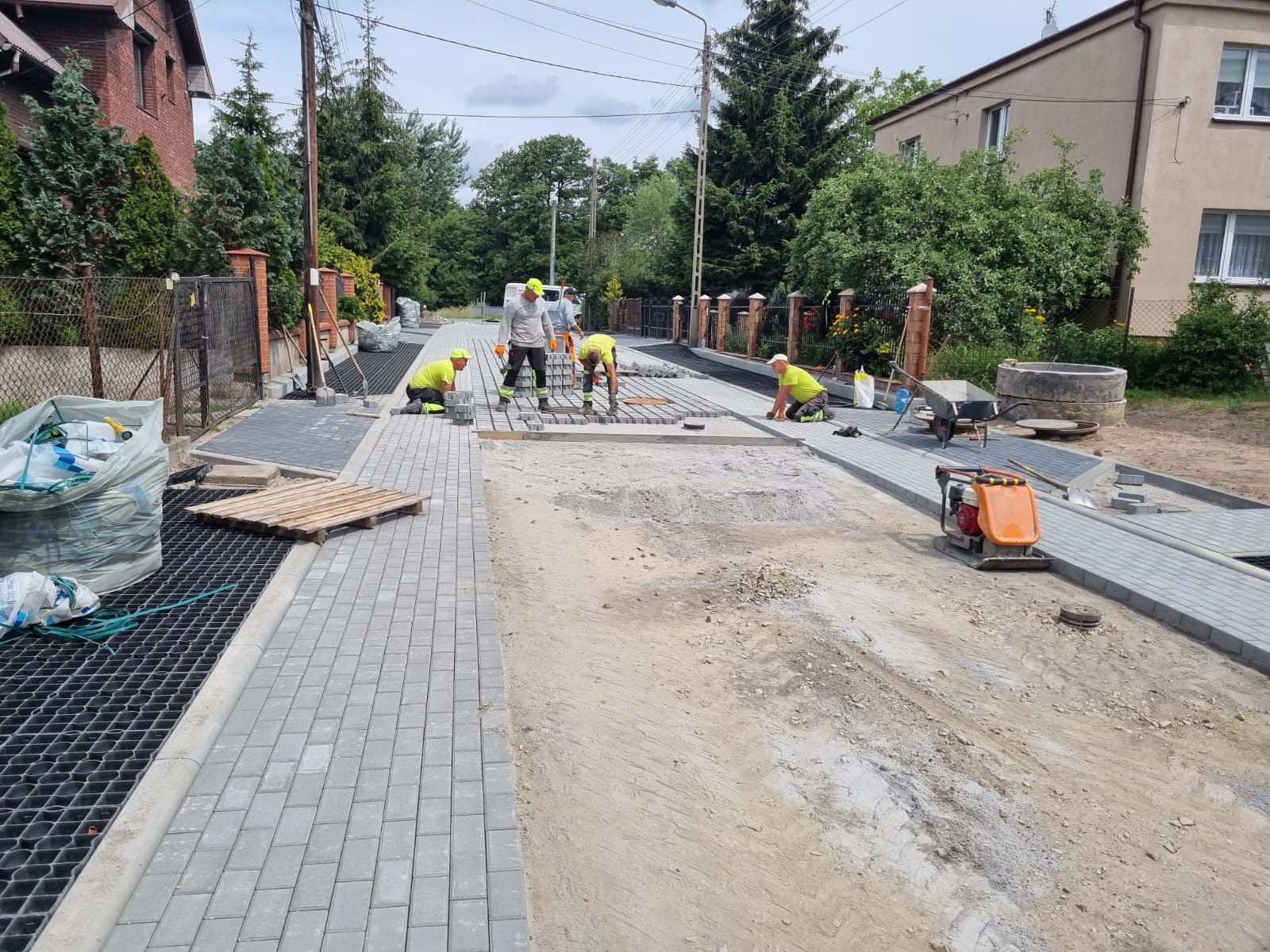 Ekipa w żółtych kamizelkach układa szarą kostkę brukową na ulicy. Część nawierzchni już gotowa, w tle domy i zieleń. Widoczna zagęszczarka i paleta z kostką.