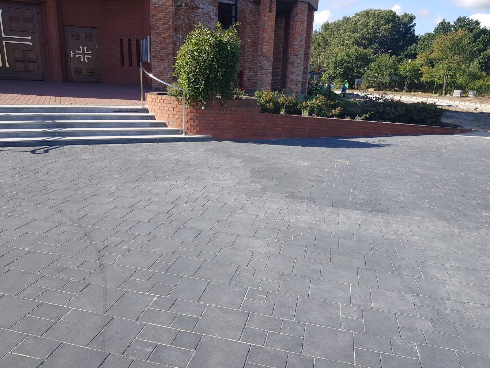 Szara kostka brukowa ułożona przed budynkiem z cegły z roślinnością. Widoczne schody z barierką i fragment terenu z drzewami w tle.