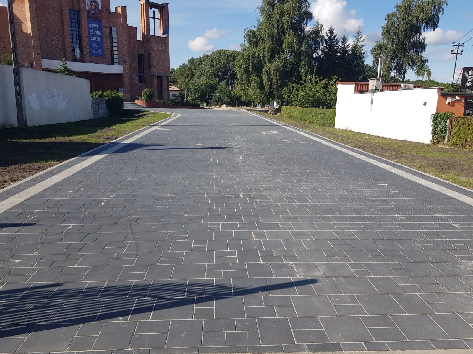 Szeroki, wybrukowany plac z ciemnej kostki z jasnymi obrzeżami, prowadzący do budynku kościoła. Widoczne drzewa i fragmenty budynków w tle, słoneczny dzień.