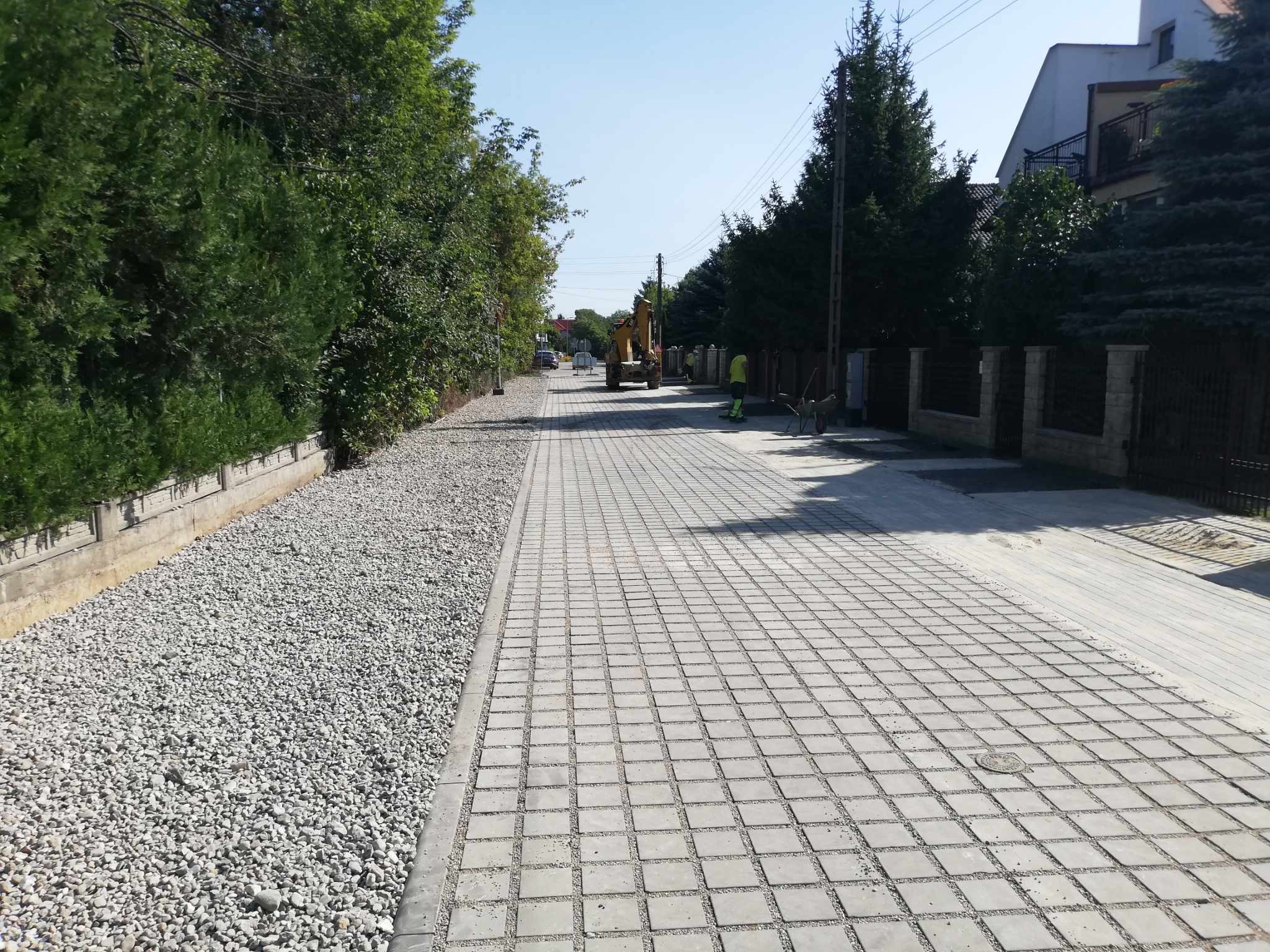 Ułożona kostka brukowa w szarym odcieniu, obok pas żwiru. W tle widoczny żółty sprzęt budowlany i pracownik w odblaskowej kamizelce. Domy i zieleń w tle.