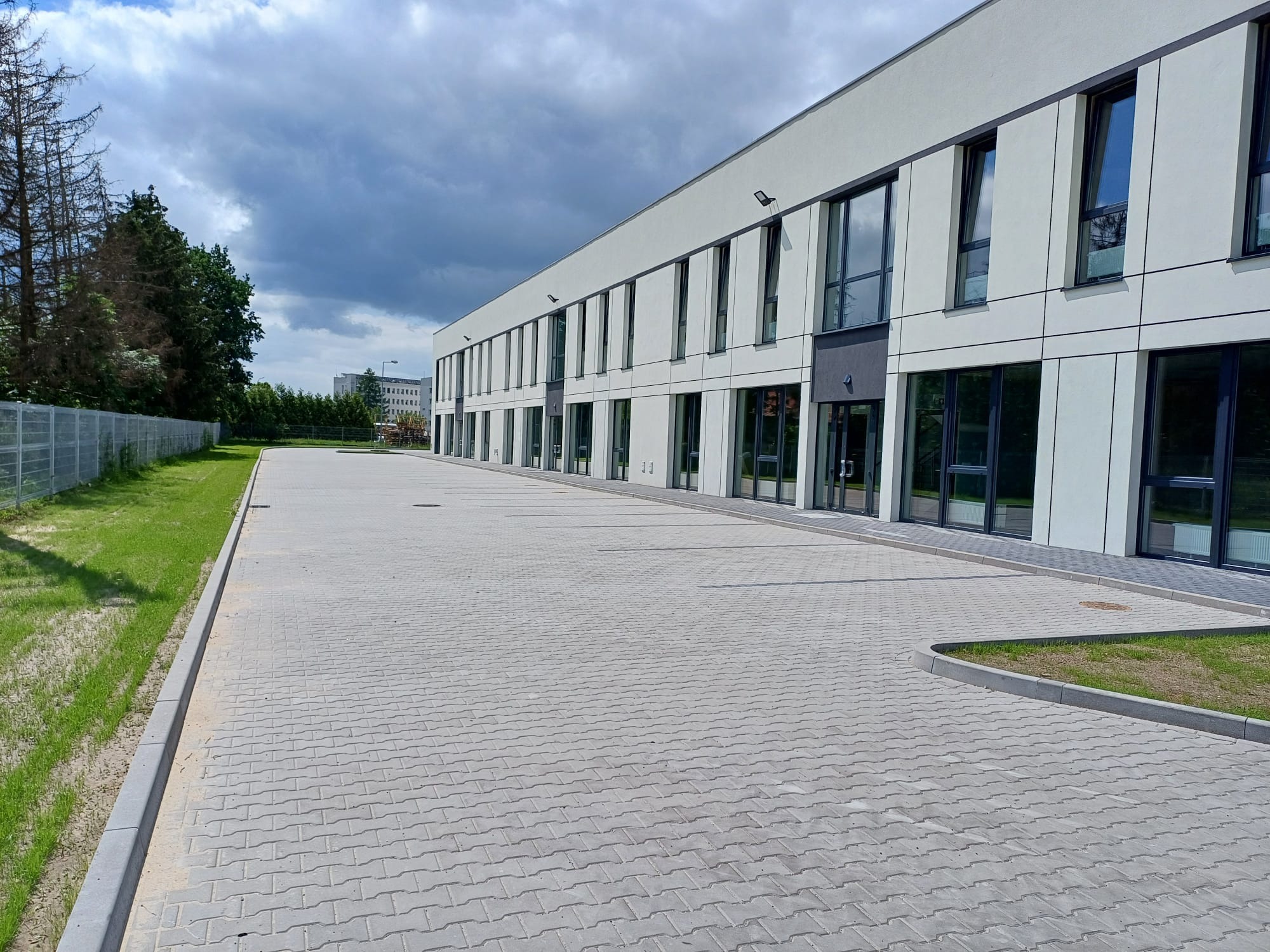 Szara kostka brukowa na parkingu przed nowoczesnym budynkiem biurowym z dużymi oknami. Widoczne trawniki i fragment ogrodzenia. Pochmurne niebo.